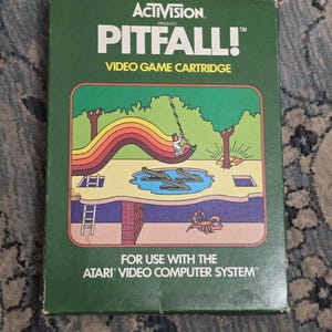 May include: A green rectangular box for the Pitfall! video game cartridge by Activision. The front features a colorful illustration of a character swinging on a vine over crocodiles, with a scorpion below ground and the Atari system text.
