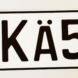 May include: A rectangular German license plate with the text "M KÄ5012". The plate features the German flag, a coat of arms, and a "D" for Deutschland. The plate has a white background with black lettering.