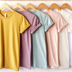 May include: A row of five short-sleeved crew neck t-shirts hanging on wooden hangers. The shirts are displayed in a color gradient from left to right, featuring mustard yellow, dusty mauve, sage green, soft blush pink, and off-white solid colors.
