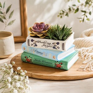 May include: A stack of three ceramic book-shaped planters. The top white planter is titled Wuthering Heights and holds two succulents. The middle light blue planter is titled Emma. The bottom green planter is titled Little Women with floral details.