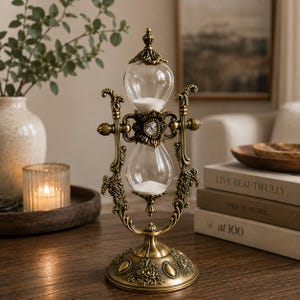 May include: An antique-style hourglass featuring a metallic bronze frame with intricate floral engravings and a central crystal accent. The clear glass bulbs contain white sand, held within a rotating structure supported by a circular base.