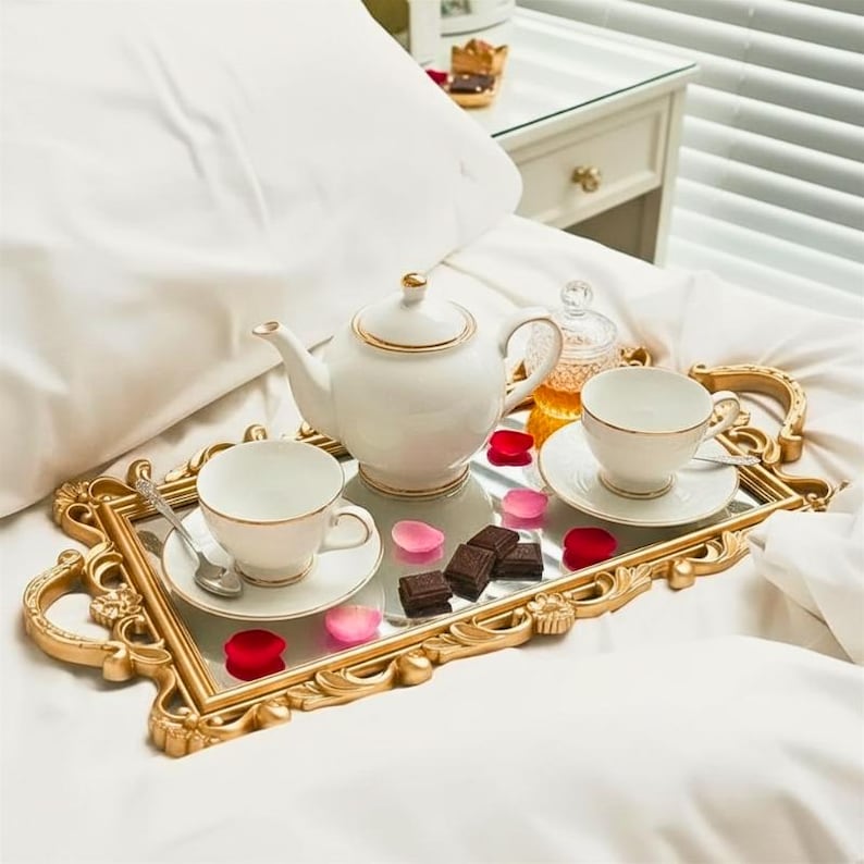 Pode incluir: Uma bandeja de servir retangular dourada com al&ccedil;as ornamentadas e base espelhada, contendo um bule de porcelana branca com borda dourada, duas x&iacute;caras combinando com pires, uma colher de prata, um pote de mel e peda&ccedil;os de chocolate amargo.