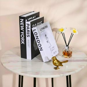 May include: A decorative display on a round table. Faux books labelled 'New York', 'Paris', and 'London' are arranged with a gold bird ornament and a diffuser with flowers. The table has a marble-effect top. The books are in a black and white color scheme.