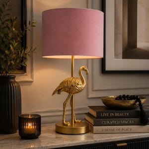 May include: A gold flamingo table lamp with a pink lampshade. The lamp is on a marble surface with books, a candle, and a decorative bowl. The books are titled "LIVE IN BEAUTY" and "CURATED SPACES".