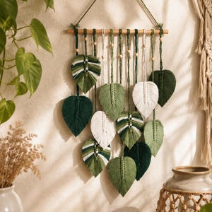 May include: A macrame wall hanging featuring multiple leaf-shaped fiber elements in shades of dark green, sage green, and white. The leaves are suspended from a horizontal wooden dowel by knotted cords, accented with small wooden beads.
