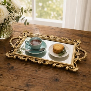 Pode incluir: Uma bandeja de servir retangular com espelho, moldura dourada ornamentada e duas al&ccedil;as laterais. A bandeja cont&eacute;m uma x&iacute;cara de ch&aacute; azul-claro sobre um pires a combinar com uma colher de prata e um pequeno prato branco com tr&ecirc;s biscoitos redondos empilhados.