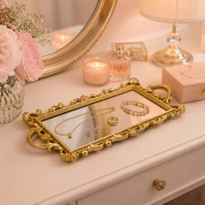 Pode incluir: Uma bandeja de toucador retangular com espelho, moldura de metal dourado ornamentada e duas pegas laterais. A bandeja cont&eacute;m um delicado colar de corrente dourada, uma pulseira de contas, um anel dourado e dois pequenos brincos.