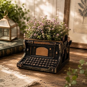 May include: A black vintage-style typewriter planter with a distressed gold finish, featuring the text NO and 3 on the front corners. The typewriter is filled with a dense cluster of small, light purple flowers and green foliage.