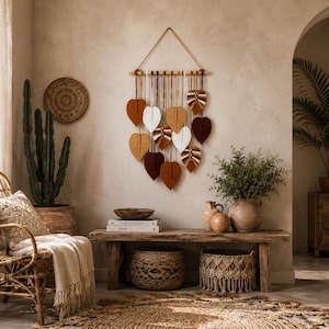 May include: A wall hanging featuring multiple macrame leaves in shades of white, tan, mustard yellow, and deep burgundy. The leaves are suspended from a horizontal wooden dowel by individual cords, creating a layered, cascading arrangement.