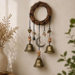 May include: A wall hanging featuring a circular brown twig wreath with five black cords suspended from it. The cords hold three brass bells, two small glass vials with cork stoppers, patterned wooden beads, and a silver crescent moon charm.