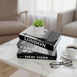 May include: Three decorative books with the city names "London", "Paris", and "New York" in white lettering. A small succulent in a white pot sits on top. A cup of coffee and glasses are on the marble table.
