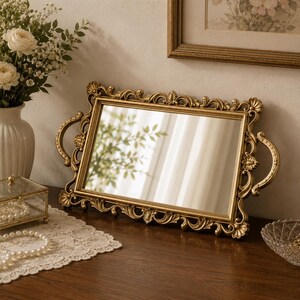 Pode incluir: Uma bandeja de espelho retangular em tom dourado com uma moldura ornamentada e curva, detalhes florais decorativos e duas al&ccedil;as laterais curvas. A superf&iacute;cie refletora &eacute; clara e a moldura tem um acabamento met&aacute;lico com detalhes em relevo.