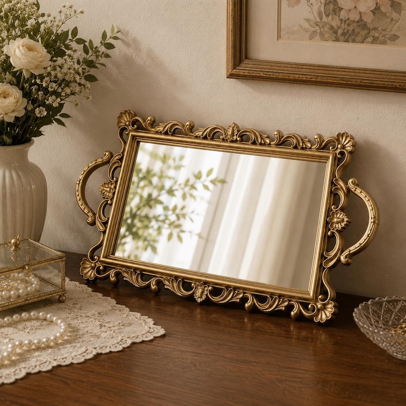 Pode incluir: Uma bandeja de espelho retangular em tom dourado com uma moldura ornamentada e curva, detalhes florais decorativos e duas al&ccedil;as laterais curvas. A superf&iacute;cie refletora &eacute; clara e a moldura tem um acabamento met&aacute;lico com detalhes em relevo.