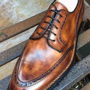 May include: A brown leather dress shoe with a classic design. The shoe features a wingtip toe, intricate stitching, and black laces. The leather has a polished finish, and the shoe is set on a dark sole.