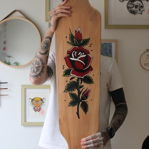 May include: A longboard skateboard with a natural wood finish. The board features a large, detailed red rose design with green leaves and stems. The skateboard is held up in front of a white wall with framed artwork.