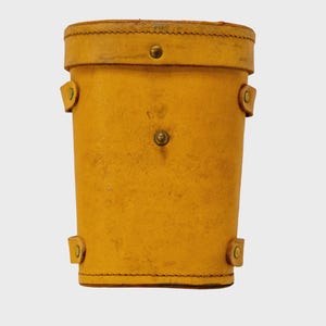 May include: A cylindrical, yellow leather container with a lid, secured by brass rivets. The container has a stitched seam around the top and bottom, and small leather loops with rivets on the sides. The overall design suggests a vintage or antique aesthetic.