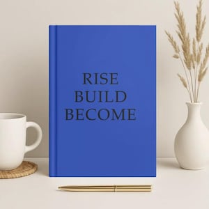 Op de afbeelding: Een blauw notitieboek met de woorden "RISE BUILD BECOME" in zwart gedrukt. Een gouden pen ligt voor het notitieboek. Een witte mok en een decoratieve vaas met gedroogde planten staan ook in de afbeelding.