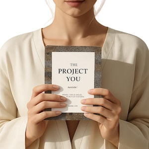 Puede incluir: Una persona sostiene un libro titulado "THE PROJECT YOU". El libro tiene una portada blanca con texto negro y un borde texturizado y multicolor. Texto adicional es visible debajo del título. La persona lleva una blusa color crema.