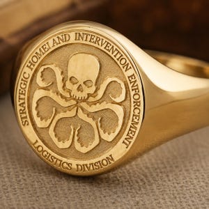 May include: A gold-toned signet ring featuring a circular face with an engraved skull and octopus-like tentacles design. The text STRATEGIC HOMELAND INTERVENTION ENFORCEMENT LOGISTICS DIVISION is embossed around the central emblem on the ring face.