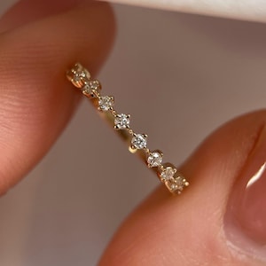 Puede incluir: Un delicado anillo de oro con una serie de pequeños diamantes brillantes. La banda del anillo es de oro fino y pulido, y los diamantes están espaciados uniformemente en la parte superior. El anillo se sostiene entre los dedos, mostrando su elegante diseño.