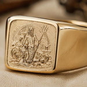 Pode incluir: Um anel de sinete em ouro com uma face quadrada apresentando uma gravação detalhada de uma figura barbuda com coroa e tridente, representada emergindo de ondas estilizadas dentro de uma borda circular numa banda de ouro polido.