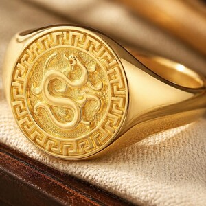 Peut inclure: Une bague chevalière en or poli présentant un plateau circulaire avec un emblème de dragon en relief au centre. Le dragon est entouré d'un motif de bordure grecque détaillé. La bague possède un anneau lisse et réfléchissant et un fond texturé.