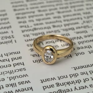 Può includere: Un anello d'oro con un diamante ovale al centro. L'anello è posto su uno sfondo di testo stampato. La fascia ha una finitura liscia e lucida.