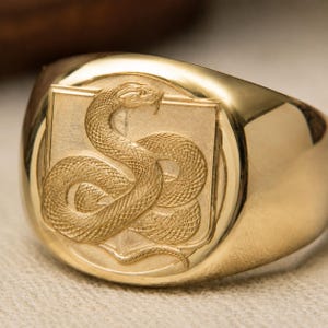 Puede incluir: Un anillo de sello de oro con una banda pulida y una cara circular plana. La cara muestra un relieve grabado detallado de una serpiente enroscada dentro de un marco cuadrado, exhibiendo texturas de escamas intrincadas y un acabado metálico.