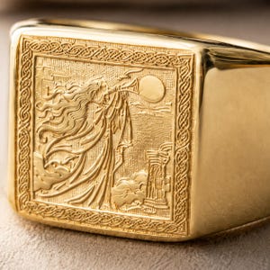 May include: A gold signet ring featuring a square face with an intricate engraved scene of a figure blowing a horn toward a circular moon. The design is framed by a detailed braided border, with a polished gold band extending from the square setting.