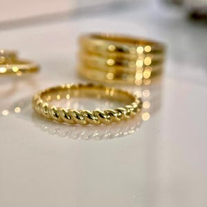 Könnte beinhalten: Nahaufnahme mehrerer goldener Ringe. Die Ringe haben unterschiedliche Designs, darunter ein gedrehter Seil-Stil und ein breiteres Band mit mehreren Schichten. Die Ringe werden auf einer weißen Oberfläche präsentiert, wobei sich das Licht im polierten Gold spiegelt.