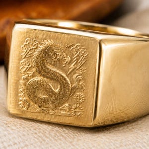 May include: A gold signet ring featuring a square face with an intricate, engraved design of a mythical dragon-like creature surrounded by stylised clouds and lightning bolts. The band is smooth and polished, showcasing a bright, reflective finish.