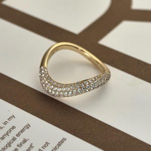 Könnte beinhalten: Ein goldfarbener Ring mit geschwungener Form. Der Ring ist mit zahlreichen kleinen, glitzernden Edelsteinen auf dem geschwungenen Teil besetzt. Der Ring liegt vor einem Hintergrund aus weißen und braunen Streifen.