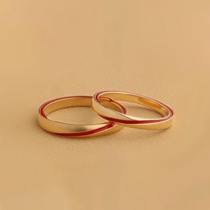 Op de afbeelding: Twee goudkleurige ringen met een rode emaille streep. De ringen worden getoond op een beige oppervlak. De ringen zijn eenvoudige banden, met een gladde, gepolijste afwerking. De rode streep voegt een vleugje kleur toe aan het verder effen ontwerp.