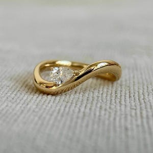 May include: A gold ring with a pear-shaped diamond set in a swirling design. The ring's band is smooth and polished, creating a continuous loop around the finger. The diamond is clear and sparkling, adding a touch of elegance.