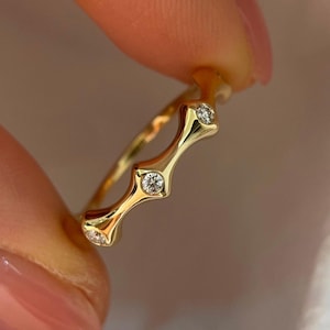 Könnte beinhalten: Ein goldener Ring mit drei kleinen, klaren Edelsteinen. Der Ring hat ein einzigartiges Design mit geschwungenen, länglichen Abschnitten zwischen den Edelsteinen. Der Ring wird zwischen zwei Fingern gehalten.