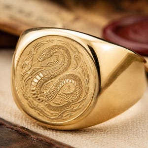 Könnte beinhalten: Ein goldener Siegelring mit einer runden Platte, die ein eingraviertes Design einer gewundenen Schlange zeigt, umgeben von stilisierten flammenartigen Mustern. Das Metall hat eine polierte, reflektierende Oberfläche, die die Details betont.