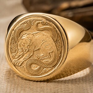 Könnte beinhalten: Ein goldener Siegelring mit einer kreisförmigen Platte, die eine detaillierte Reliefgravur eines Tigers und einer Schlange zeigt. Der polierte Goldring hat eine glatte, abgerundete Oberfläche, die sich zur Rückseite hin verjüngt.