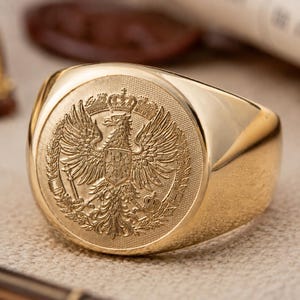 May include: A polished gold signet ring featuring a detailed, circular engraved emblem of a double-headed eagle with a crown and shield. The design includes intricate textures and fine lines, showcasing a regal heraldic motif on a smooth band.