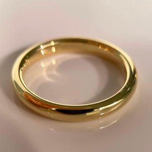 May include: A gold ring with a polished, reflective surface. The ring has a rounded profile and a simple, unadorned design. The ring is likely made of gold or a gold-coloured metal. A piece of jewellery.