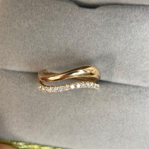 Puede incluir: Dos anillos de oro. El anillo superior tiene un diseño liso y ondulado. El anillo inferior es una banda de pequeños diamantes brillantes. Los anillos están sobre un fondo gris.