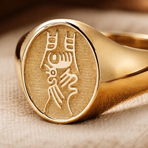 Puede incluir: Un anillo de sello de oro con una cara ovalada de fondo texturizado y un carácter chino pulido en relieve grabado en el centro. El anillo tiene una banda lisa y redondeada con un acabado brillante que refleja la luz en toda su superficie.