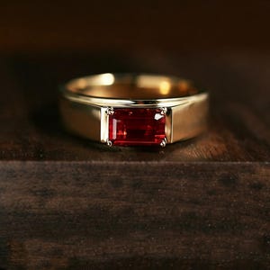 May include: A gold ring featuring a rectangular, emerald-cut red gemstone set horizontally in the center. The band is smooth, polished, and solid gold, with a wide, flat profile that tapers slightly toward the back of the ring.