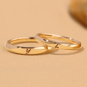 Puede incluir: Dos anillos dorados con un acabado pulido. Un anillo tiene la letra "y" grabada, y el otro la letra "L". Los anillos tienen un diseño facetado en los bordes exteriores y una banda interior lisa y redondeada. Los anillos son probablemente para una pareja.