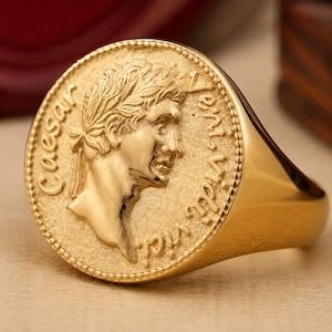 May include: A gold signet ring featuring a circular face with a raised profile of Julius Caesar wearing a laurel wreath. The text Caesar Veni Vidi Vici is embossed around the perimeter of the coin-style design, which has a textured background.