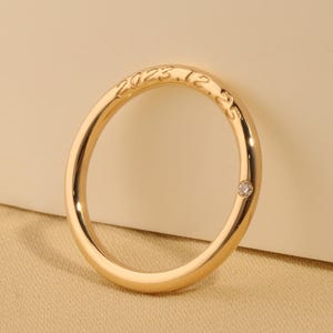 May include: A gold ring with a small diamond set into the band. The ring has the engraved text "2023.12" on the outer surface. The ring is displayed on a neutral background.
