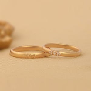 Könnte beinhalten: Zwei goldene Eheringe. Einer der Ringe hat ein Blumenmuster, der andere ist mit kleinen, funkelnden Steinen besetzt. Die Ringe sind vor einem neutralen Hintergrund platziert, was ihre elegante Schlichtheit unterstreicht. Diese Ringe symbolisieren Engagement und Liebe.