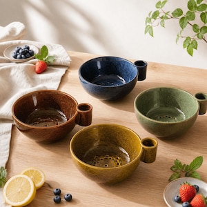 Puede incluir: Cuatro cuencos de cerámica para bayas en azul oscuro, marrón, verde oliva y amarillo mostaza. Cada cuenco cuenta con un asa lateral, un pequeño soporte cilíndrico y una base perforada para el drenaje. Los cuencos están dispuestos sobre una superficie de madera con arándanos, rodajas de limón y fresas esparcidas.
