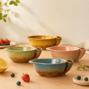 May include: A set of four ceramic bowls in yellow, green, pink, and blue, each featuring a side handle, a pouring spout, and a textured brown base. The bowls are arranged on a surface with scattered strawberries, blueberries, and a lemon slice.