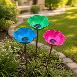 Puede incluir: Tres estacas de jardín con grandes flores brillantes en colores verde vibrante, azul intenso y rosa fuerte. Cada flor presenta un centro oscuro y texturizado, montada sobre un tallo metálico delgado de color marrón para su uso en el suelo.