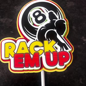 Op de afbeelding: Een kleurrijke taarttopper met de tekst "RACK EM UP" in geel en rood, met een zwart-wit 8-ball ontwerp en een silhouet van een persoon die pool speelt. De topper staat op een witte stok.